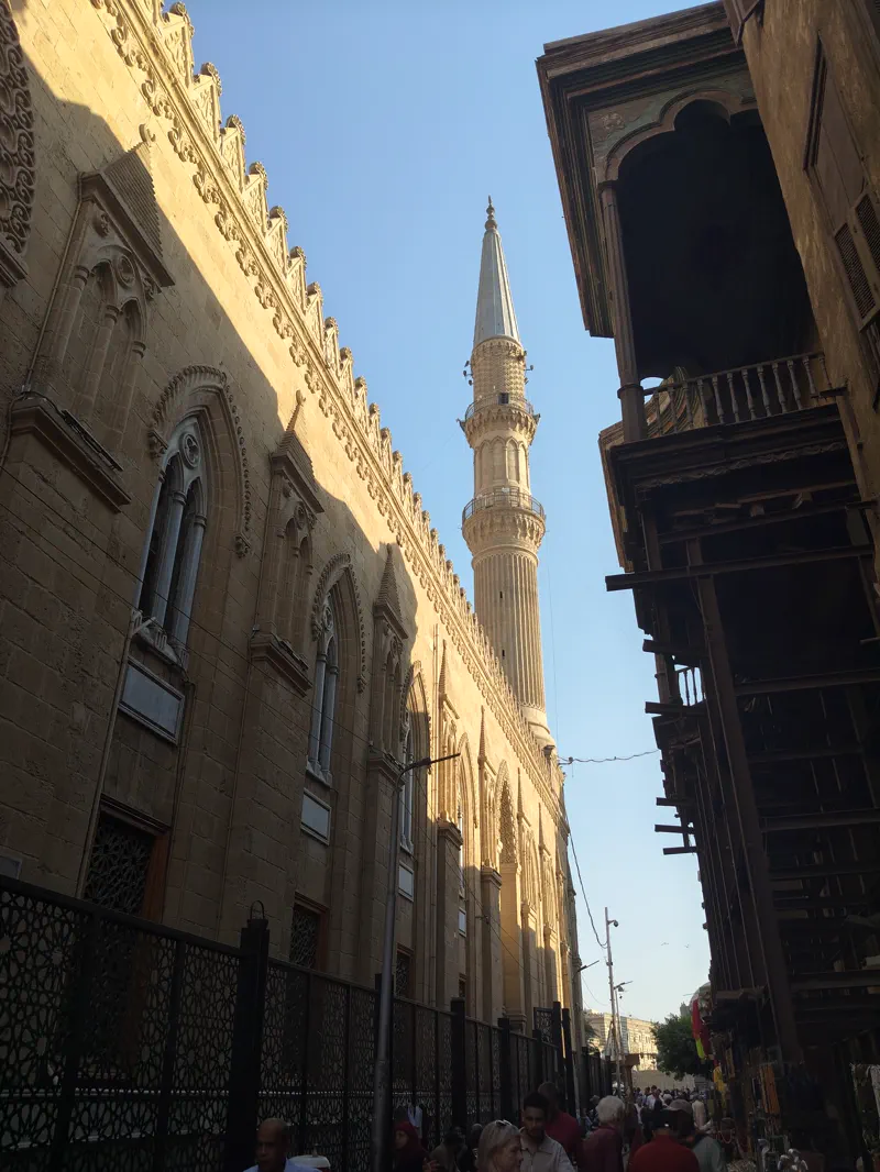 Old Cairo lane with a historic stone facade, wooden balconies, and a minaret